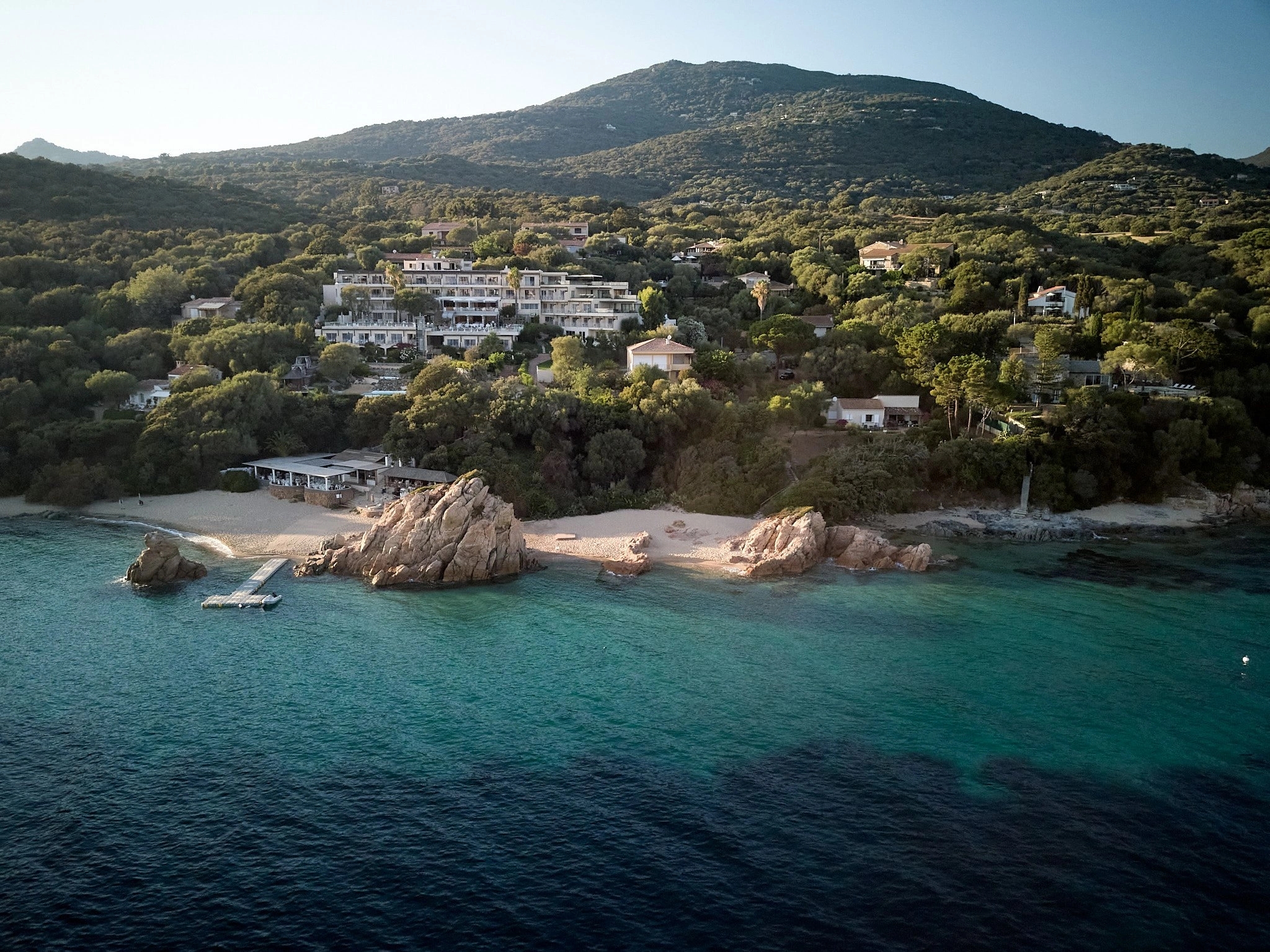 Vue aérienne d'une plage méditerranéenne avec rochers, villa luxueuse et montagne boisée en arrière-plan