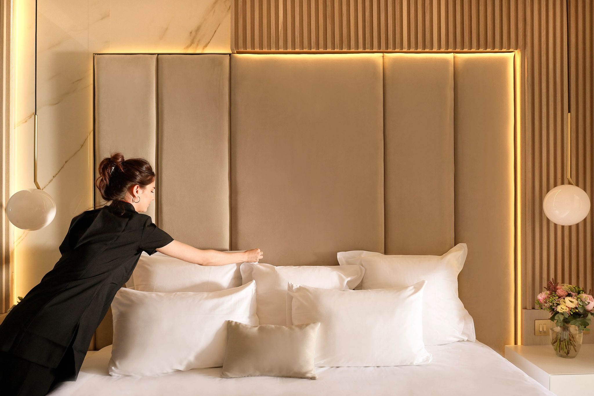 Femme en uniforme noir arrangeant les oreillers blancs sur un lit avec tête de lit rembourrée beige et éclairage doré